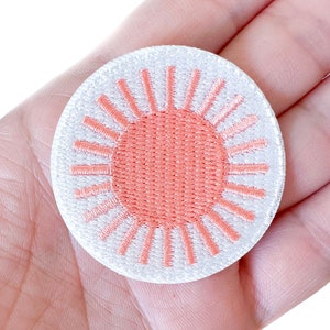 May include: A white embroidered patch with a pink sun design. The sun has a circle in the center and rays radiating out from the center.