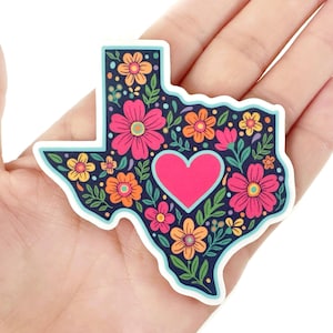 May include: A sticker in the shape of Texas, filled with colorful flowers and a pink heart. The background is navy blue, and the flowers are pink, orange, and yellow. The sticker has a white border.