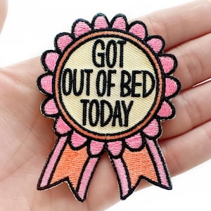 May include: A round, embroidered patch with the text "GOT OUT OF BED TODAY" in black capital letters. The patch has a light yellow center, a pink scalloped border, and pink and orange ribbons. The patch is held in a hand.