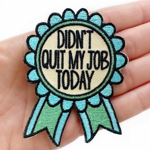 May include: Embroidered patch in the shape of a teal and green award ribbon. The patch has the text "DIDN'T QUIT MY JOB TODAY" in black lettering on a beige background. The ribbon has a scalloped edge.