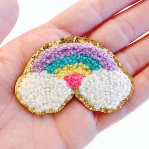 May include: A rainbow-colored felt patch with a gold glitter border. The rainbow has purple, blue, green, yellow, and pink stripes. The rainbow is surrounded by two white fluffy clouds.