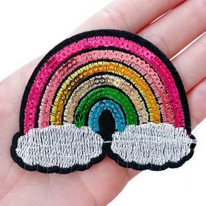 May include: A colorful rainbow patch with a black border and white clouds. The rainbow features pink, orange, yellow, green, and blue sequins. This embroidered patch is suitable for clothing, bags, or other fabric items.