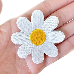 May include: A white daisy flower patch with a yellow center. The patch is embroidered with a textured appearance. The daisy is held in a hand, showcasing the size and detail of the floral design. This patch is suitable for clothing and accessories.