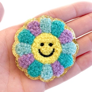 May include: A yellow smiley face surrounded by a flower made of purple, blue, and green fuzzy fabric. The flower is outlined in gold glitter.