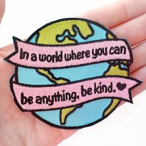 May include: Embroidered patch featuring a light blue globe with green continents. A pink banner wraps around the globe, displaying the text "In a world where you can be anything, be kind." A black heart is on the right side of the banner.
