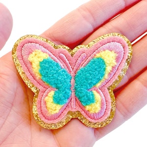May include: A colorful butterfly-shaped applique with a pink, yellow, and teal design. The butterfly has a gold glitter border and is made of felt. This decorative applique can be used for crafts and embellishments.