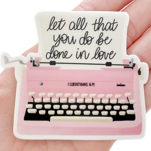 May include: A pink typewriter sticker with the text "let all that you do be done in love" in black script. The sticker also includes the Bible verse "I Corinthians 16:14". The typewriter has white keys and black accents.