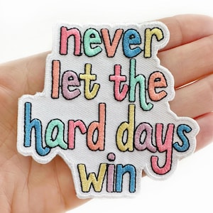 May include: Embroidered patch with the text "never let the hard days win" in colorful letters. The patch has a white background and a black border. The patch is held in a hand.