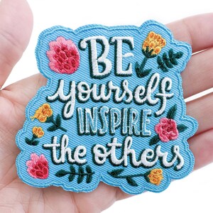 May include: Blue embroidered iron-on patch with white text that reads "Be yourself inspire the others" surrounded by colorful flowers.