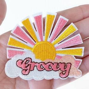 May include: Embroidered patch featuring a retro sun design with yellow and pink rays, a yellow sun, and a cloud with the word "Groovy" in pink. The patch has a white border.