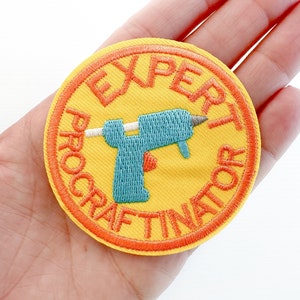 May include: A round, yellow embroidered patch with the words "EXPERT PROCRAFTINATOR" in orange. A blue glue gun is in the center. The patch has an orange border.