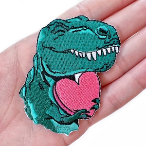 May include: Green embroidered dinosaur holding a pink heart. The dinosaur has a toothy grin and is holding the heart in its front paws.