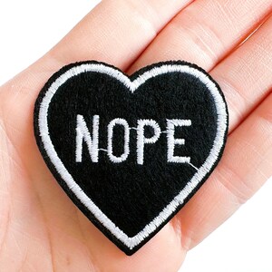 May include: Black heart-shaped patch with white embroidered border and the word "NOPE" in white block letters. The patch is made of a felt-like material and is held in a hand.