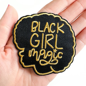 May include: Black patch with gold embroidered text that reads "BLACK GIRL magic". The patch has a black background with a gold border. The patch is round with a wavy edge.