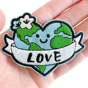 May include: Embroidered patch featuring a smiling Earth in a heart shape, with green continents and blue oceans. A banner below reads "LOVE" in black letters. A white flower and black outline complete the design. Ideal for jackets, bags, or other fabric items.
