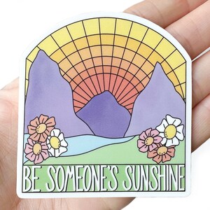 May include: A sticker with a retro-style illustration of mountains, a sunset, and flowers. The text "BE SOMEONE'S SUNSHINE" is printed in white on a green background.