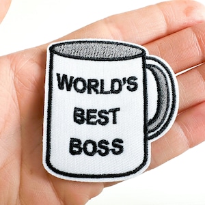 May include: Embroidered patch in the shape of a coffee mug. The white patch has a black border and a black handle. The text "WORLD'S BEST BOSS" is embroidered in black on the front of the mug.