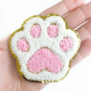 May include: A white and pink fuzzy paw print with a gold glitter outline. The paw print is a cute and whimsical design.