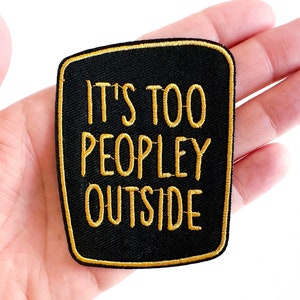 May include: Black iron-on patch with gold embroidered text that reads "It's too peopley outside".