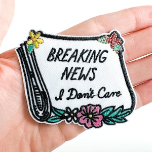 May include: Embroidered patch with the text "BREAKING NEWS I Don't Care" in black script on a white background. The patch is shaped like a newspaper and decorated with floral accents in pink, yellow, and teal. The patch is held in a hand.