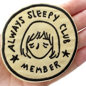 May include: A beige fabric patch with black embroidered text that reads "Always Sleepy Club Member" and a cartoon face with closed eyes.
