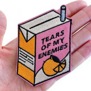 May include: Pink and orange embroidered iron-on patch shaped like a juice box with the text "Tears of my enemies" and a cartoon orange.