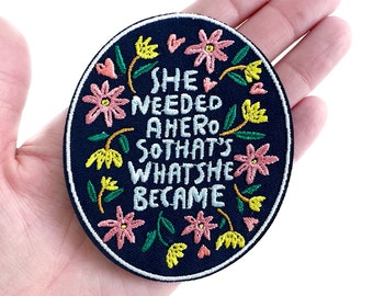 May include: A black embroidered patch with a floral border and the text "She needed a hero so that's what she became."