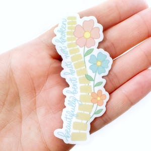 Pode incluir: Um autocolante com o texto "beautifully bent but not broken" em fonte cursiva. O autocolante apresenta um desenho de tijolos amarelos e tr&ecirc;s flores em tons de p&ecirc;ssego, azul e laranja. O autocolante &eacute; segurado numa m&atilde;o.