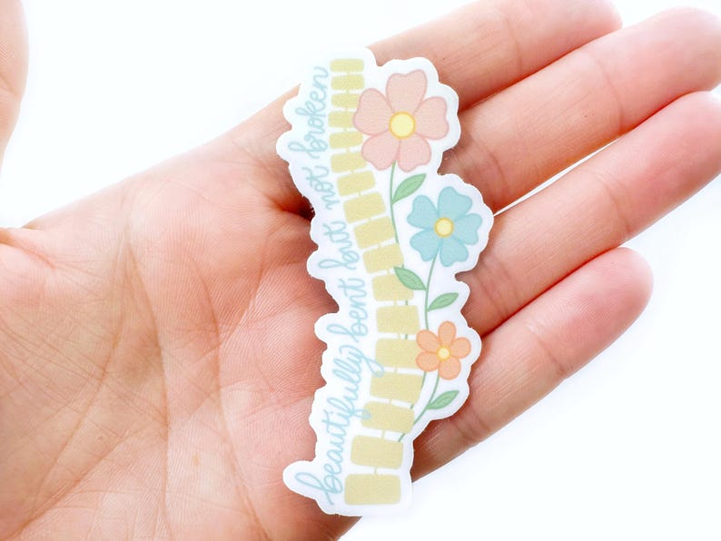 Pode incluir: Um autocolante com o texto "beautifully bent but not broken" em fonte cursiva. O autocolante apresenta um desenho de tijolos amarelos e tr&ecirc;s flores em tons de p&ecirc;ssego, azul e laranja. O autocolante &eacute; segurado numa m&atilde;o.