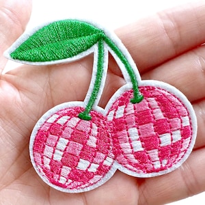 May include: Embroidered patch featuring two cherries with a disco ball design. The cherries are pink and white, with green stems and a green leaf. The patch has a white border.