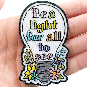 May include: White embroidered iron-on patch shaped like a light bulb with the text "Be a light for all to see" and colorful flowers around the base.