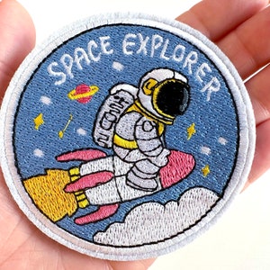 May include: Embroidered patch with a space theme. The patch features an astronaut riding a rocket ship against a blue background with the words "SPACE EXPLORER". The design includes stars, planets, and clouds.