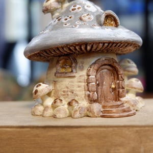 May include: A decorative mushroom-shaped house figurine featuring a grey cap with white spots, a beige textured base, a brown arched door with a gold knob, a small window, and several smaller mushroom accents surrounding the base of the structure.