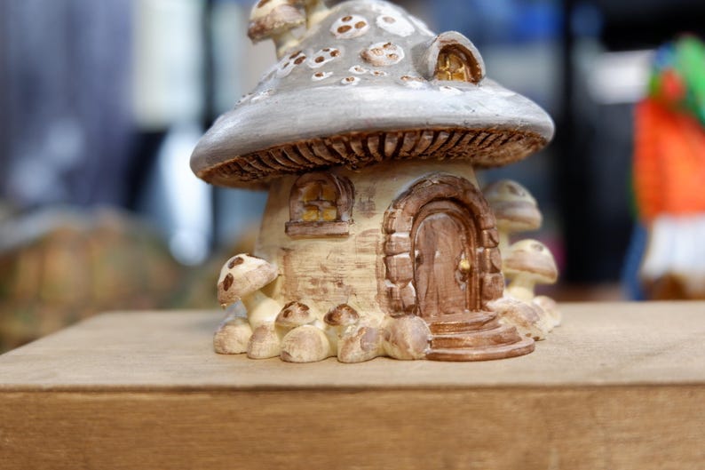 May include: A decorative mushroom-shaped house figurine featuring a grey cap with white spots, a beige textured base, a brown arched door with a gold knob, a small window, and several smaller mushroom accents surrounding the base of the structure.