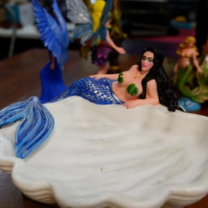 May include: A white ceramic shell-shaped dish featuring a reclining mermaid with long black hair, a blue and silver scale tail, and green leaf-shaped accents on the chest. The mermaid's tail extends over the edge of the white scalloped bowl.