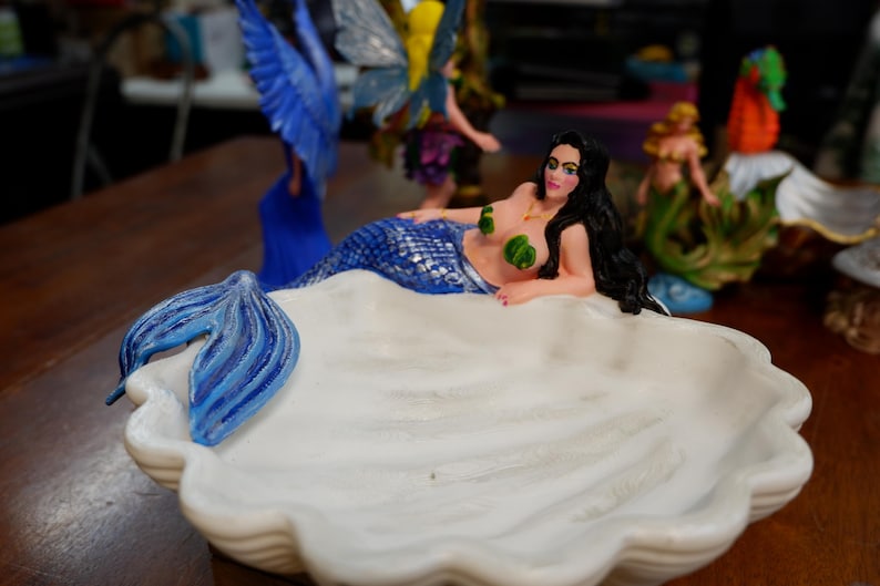 May include: A white ceramic shell-shaped dish featuring a reclining mermaid with long black hair, a blue and silver scale tail, and green leaf-shaped accents on the chest. The mermaid's tail extends over the edge of the white scalloped bowl.
