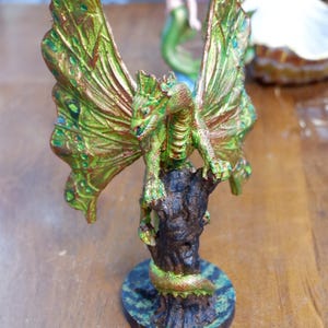 Op de afbeelding: Een geschilderd drakenbeeldje met grote, iriserende groen en koperkleurige vlinderachtige vleugels, zittend op een donkerbruine, getextureerde boomstronkvoet. De draak heeft een schilferig groen lichaam met koperkleurige accenten op een ronde voet.