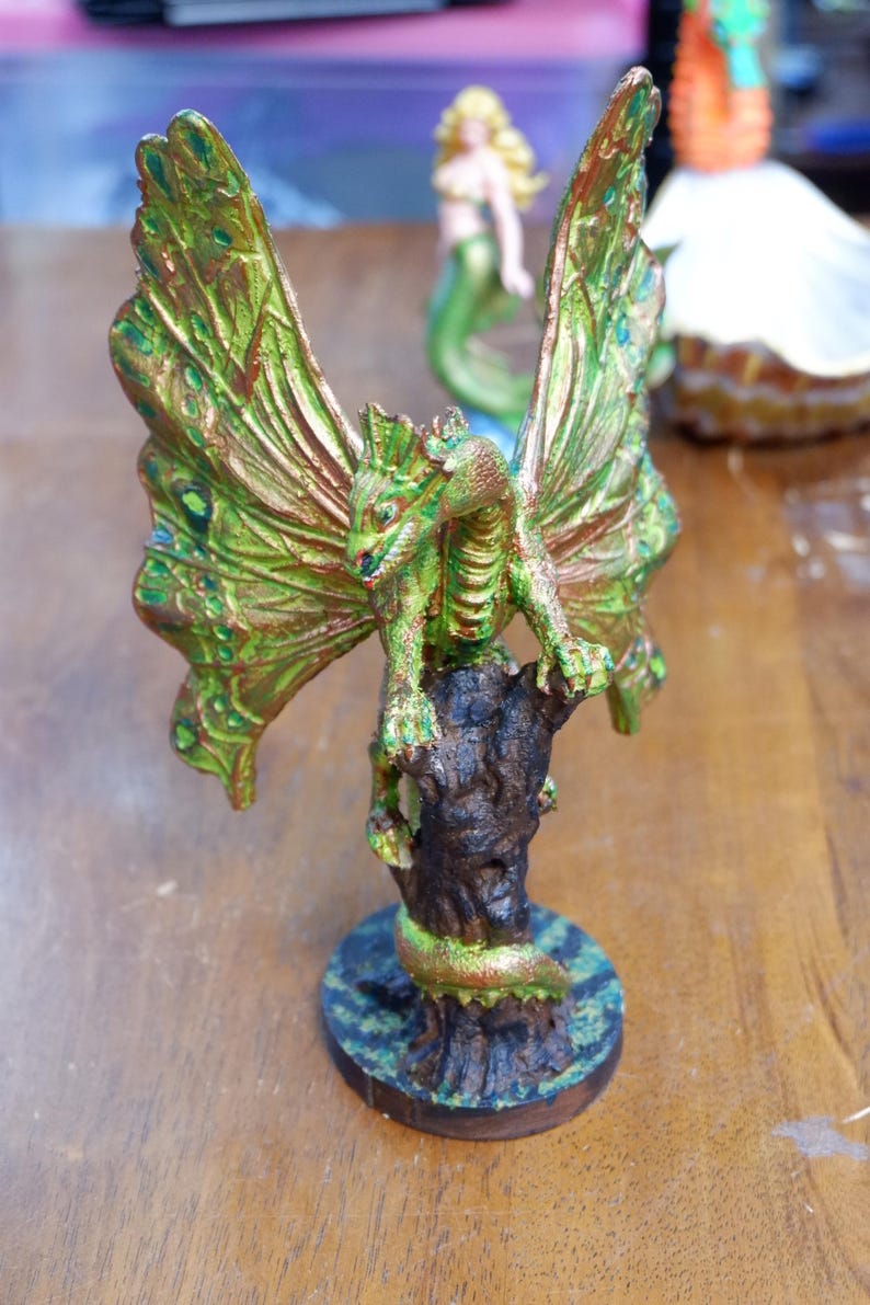Op de afbeelding: Een geschilderd drakenbeeldje met grote, iriserende groen en koperkleurige vlinderachtige vleugels, zittend op een donkerbruine, getextureerde boomstronkvoet. De draak heeft een schilferig groen lichaam met koperkleurige accenten op een ronde voet.