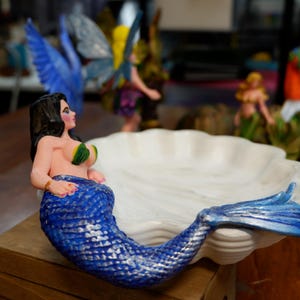 May include: A decorative dish shaped like a white scalloped seashell featuring a mermaid figure attached to the side. The mermaid has long black hair, green shell-like accents on the chest, and a textured blue tail that extends along the edge.