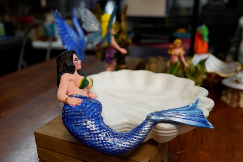 May include: A decorative dish shaped like a white scalloped seashell featuring a mermaid figure attached to the side. The mermaid has long black hair, green shell-like accents on the chest, and a textured blue tail that extends along the edge.