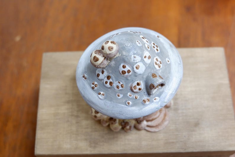 May include: A mushroom-shaped figurine with a light gray cap featuring white and brown circular spots. The top of the cap has two small protruding mushroom accents and a small arched opening. The base is textured with brown and tan circular details.