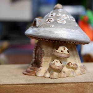May include: A decorative mushroom-shaped house figurine featuring a large, silver-grey cap with white spots. The base is cream-colored with a small arched doorway and several smaller, brown-spotted mushroom accents clustered near the entrance.