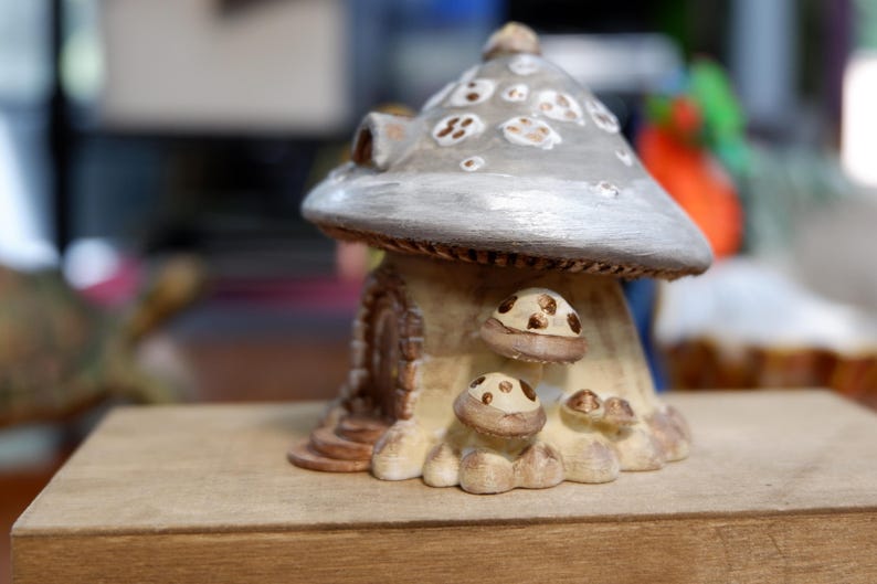 May include: A decorative mushroom-shaped house figurine featuring a large, silver-grey cap with white spots. The base is cream-colored with a small arched doorway and several smaller, brown-spotted mushroom accents clustered near the entrance.