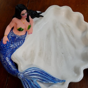 May include: A white seashell-shaped dish featuring a sculpted mermaid with long black hair, green leaf-shaped top, and a textured blue tail. The mermaid is positioned along the curved edge of the white, scalloped shell bowl, which has a smooth interior.