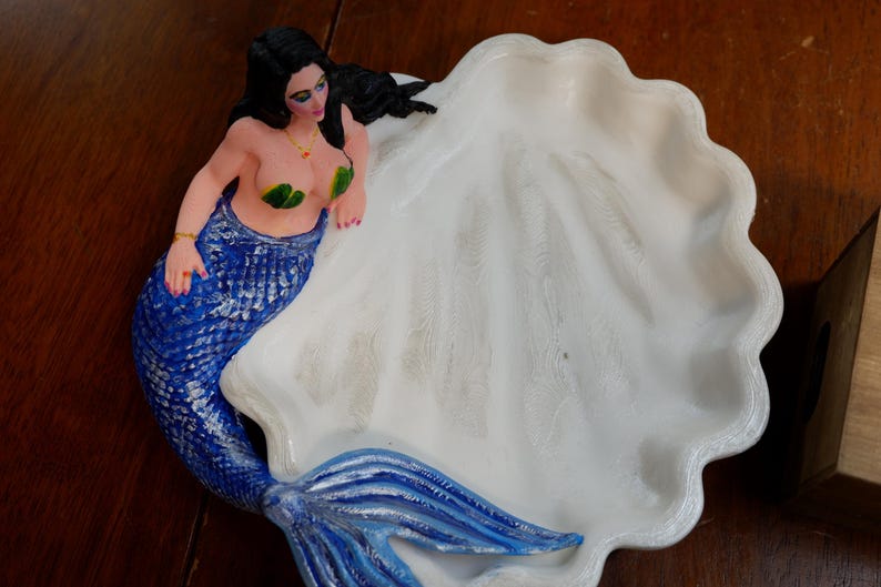 May include: A white seashell-shaped dish featuring a sculpted mermaid with long black hair, green leaf-shaped top, and a textured blue tail. The mermaid is positioned along the curved edge of the white, scalloped shell bowl, which has a smooth interior.