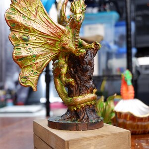 Op de afbeelding: Een drakenbeeldje met iriserende groene en koperkleurige schubben, grote getextureerde vleugels en een lange staart die om een donkerbruine boomstronkbasis is gewikkeld. Het wezen staat op een rechthoekig houten blok met een ovale uitsparing.