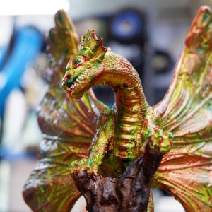 Op de afbeelding: Een drakenfiguur met getextureerde schubben in tinten groen, koper en goud. Het wezen heeft grote, uitgestrekte vleugels en zit bovenop een donkere, ruwe, boomachtige structuur. De kop is opzij gedraaid en toont scherpe tanden.