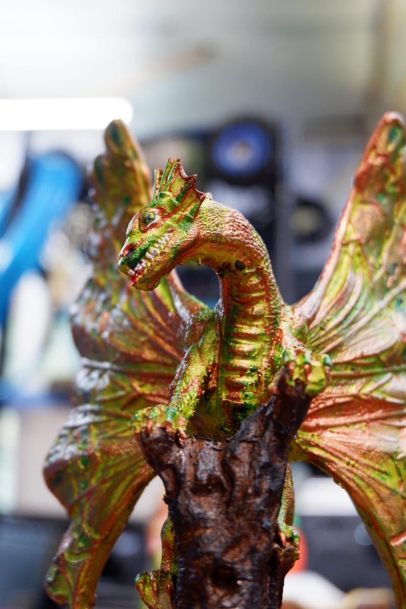 Op de afbeelding: Een drakenfiguur met getextureerde schubben in tinten groen, koper en goud. Het wezen heeft grote, uitgestrekte vleugels en zit bovenop een donkere, ruwe, boomachtige structuur. De kop is opzij gedraaid en toont scherpe tanden.