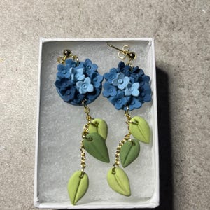 K&ouml;nnte beinhalten: Ein Paar blaue und gr&uuml;ne florale Ohrringe mit goldenen Ketten. Die Ohrringe befinden sich in einer wei&szlig;en Schachtel.