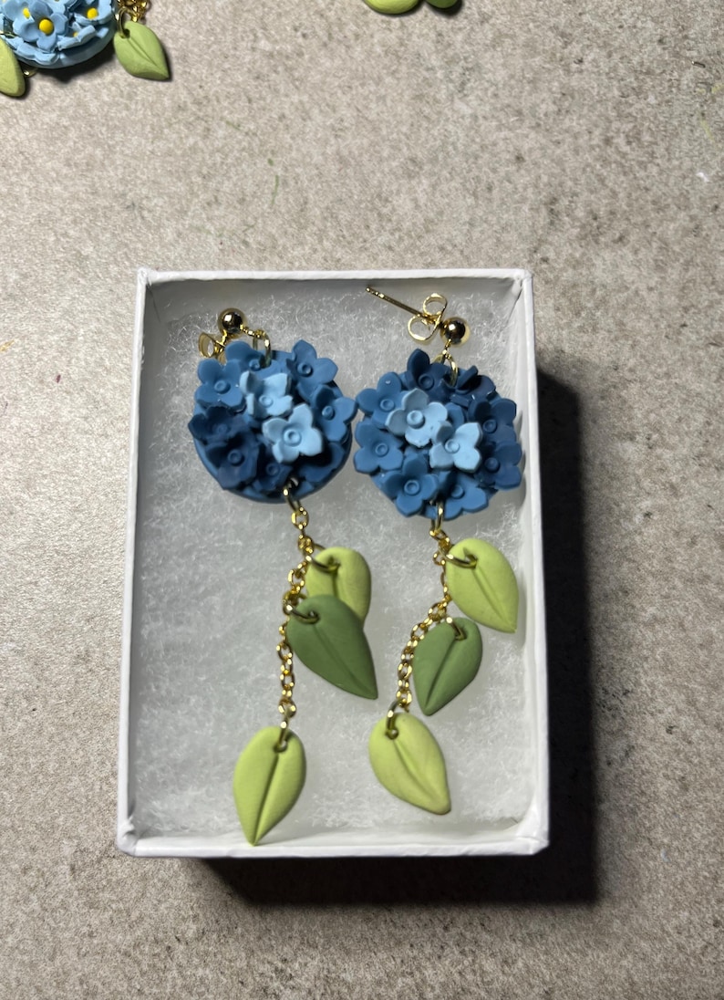 K&ouml;nnte beinhalten: Ein Paar blaue und gr&uuml;ne florale Ohrringe mit goldenen Ketten. Die Ohrringe befinden sich in einer wei&szlig;en Schachtel.